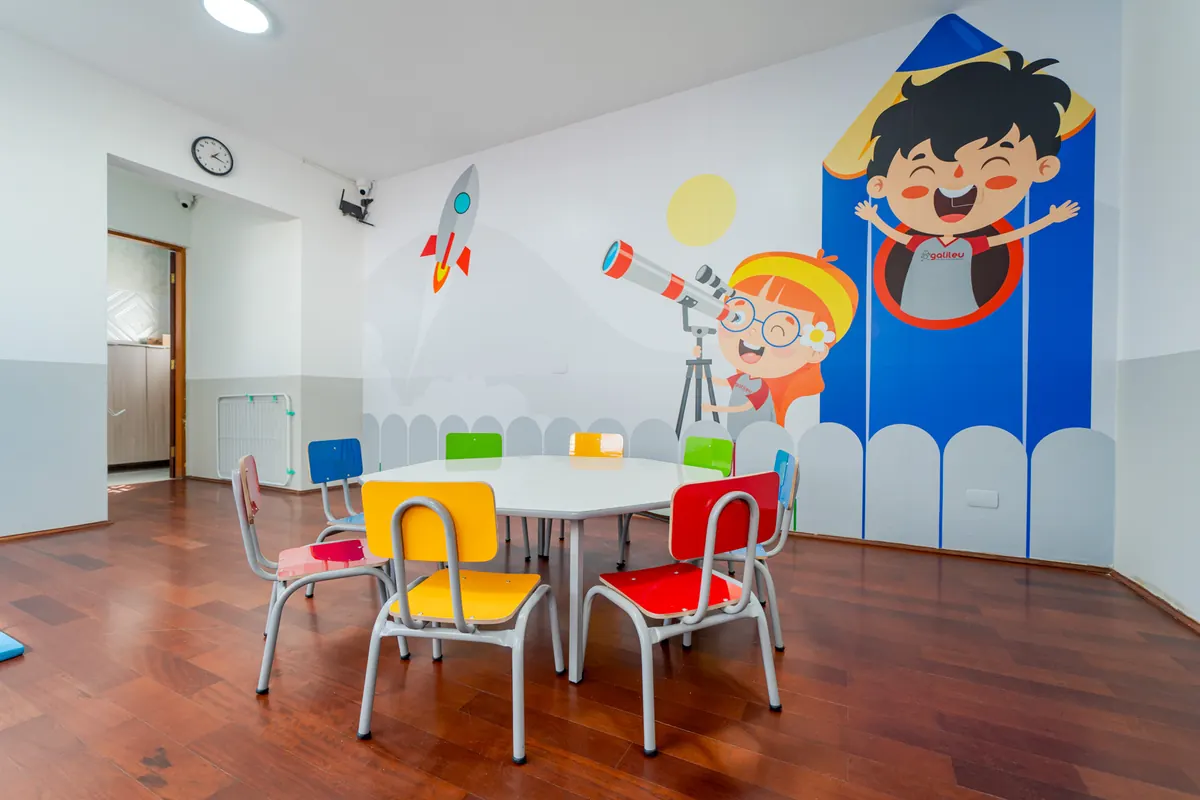 Sala de aula com mesa redonda, cadeirinhas coloridas e mural de astronauta