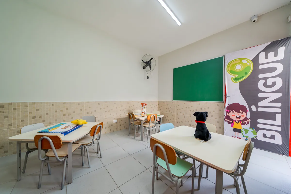 Sala bilíngue com mesas, cadeiras coloridas, lousa verde e banner Bilíngue